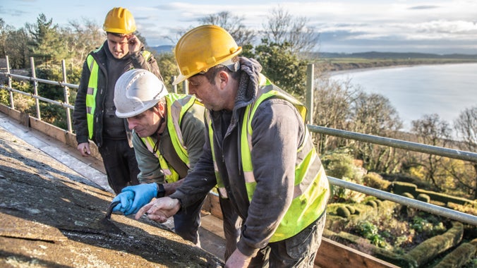 Close-up of a slate roof with builders, scaffolding and a stone chimney under renovation